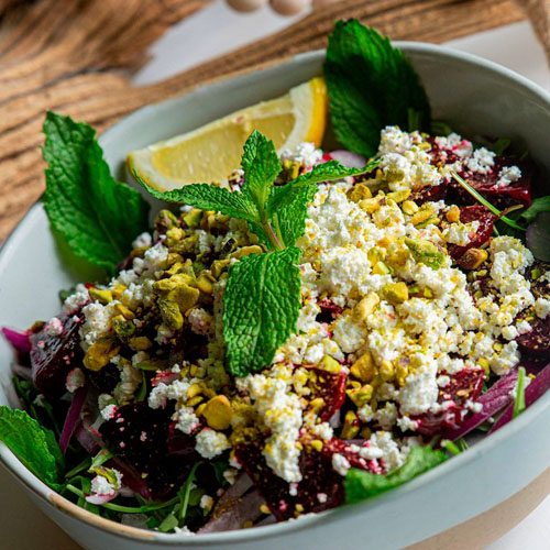 Bowl of granola topped with banana slices, blueberries, strawberries, shredded coconut, and fresh mint leaves with a wooden spoon beside it.