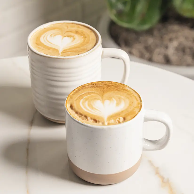 Two women wearing sunglasses sitting at a wooden table outdoors, enjoying a meal with plates of food and drinks, surrounded by large green plants.