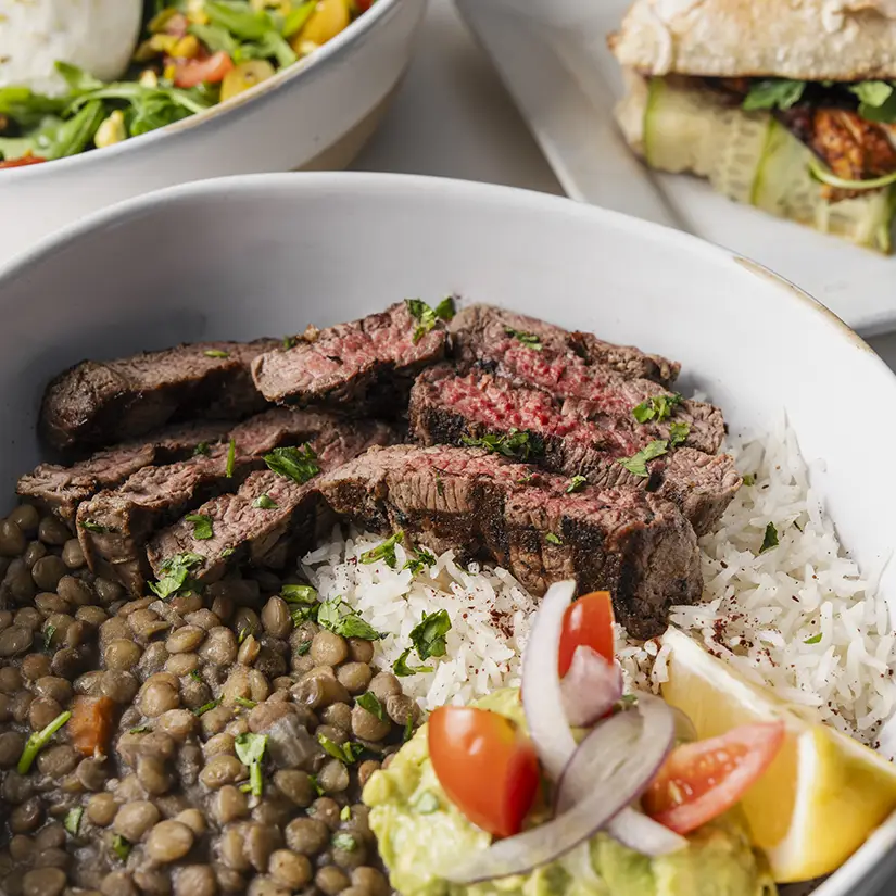 Cast iron skillet with eggs in spicy tomato sauce topped with avocado slices, served with pita bread and two dipping sauces on a tray, alongside an orange juice drink.