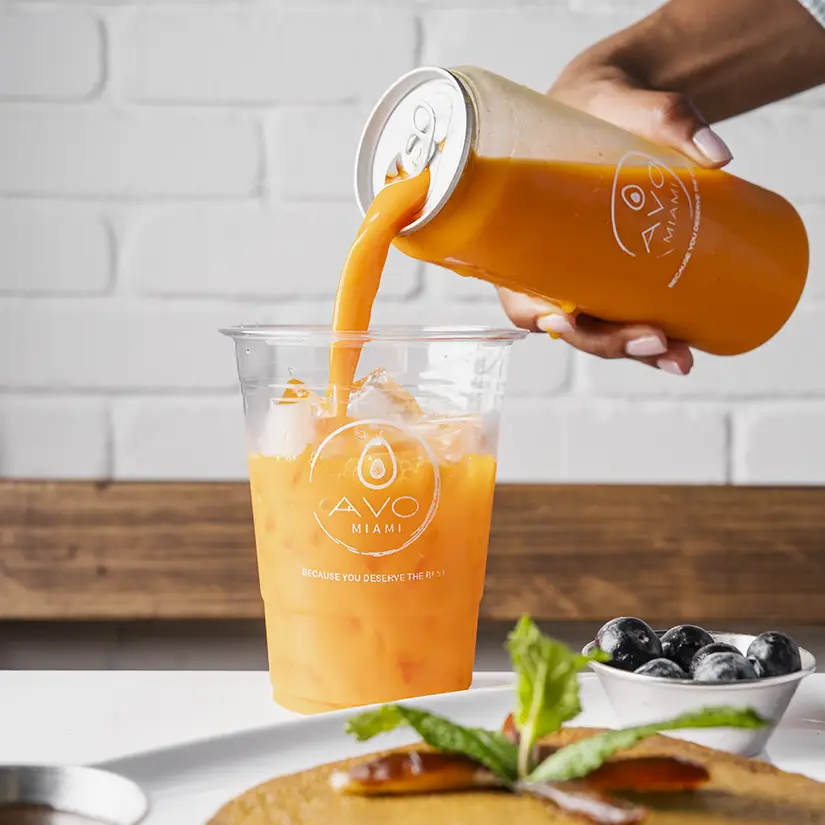 Top view of a wooden table with plates of omelettes, avocado pancakes with berries, toast, and three hands holding different beverages including a latte, iced drink with chopsticks, and a green smoothie garnished with mint.