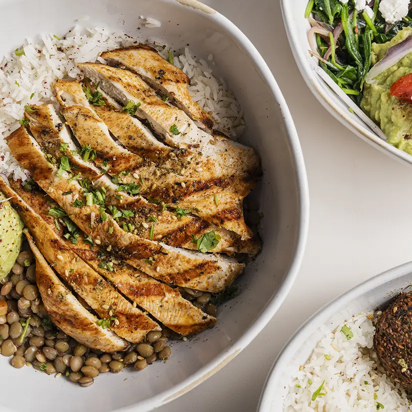 Outdoor café seating with wooden chairs, white tables, large potted green plants, and fringed beige umbrellas.