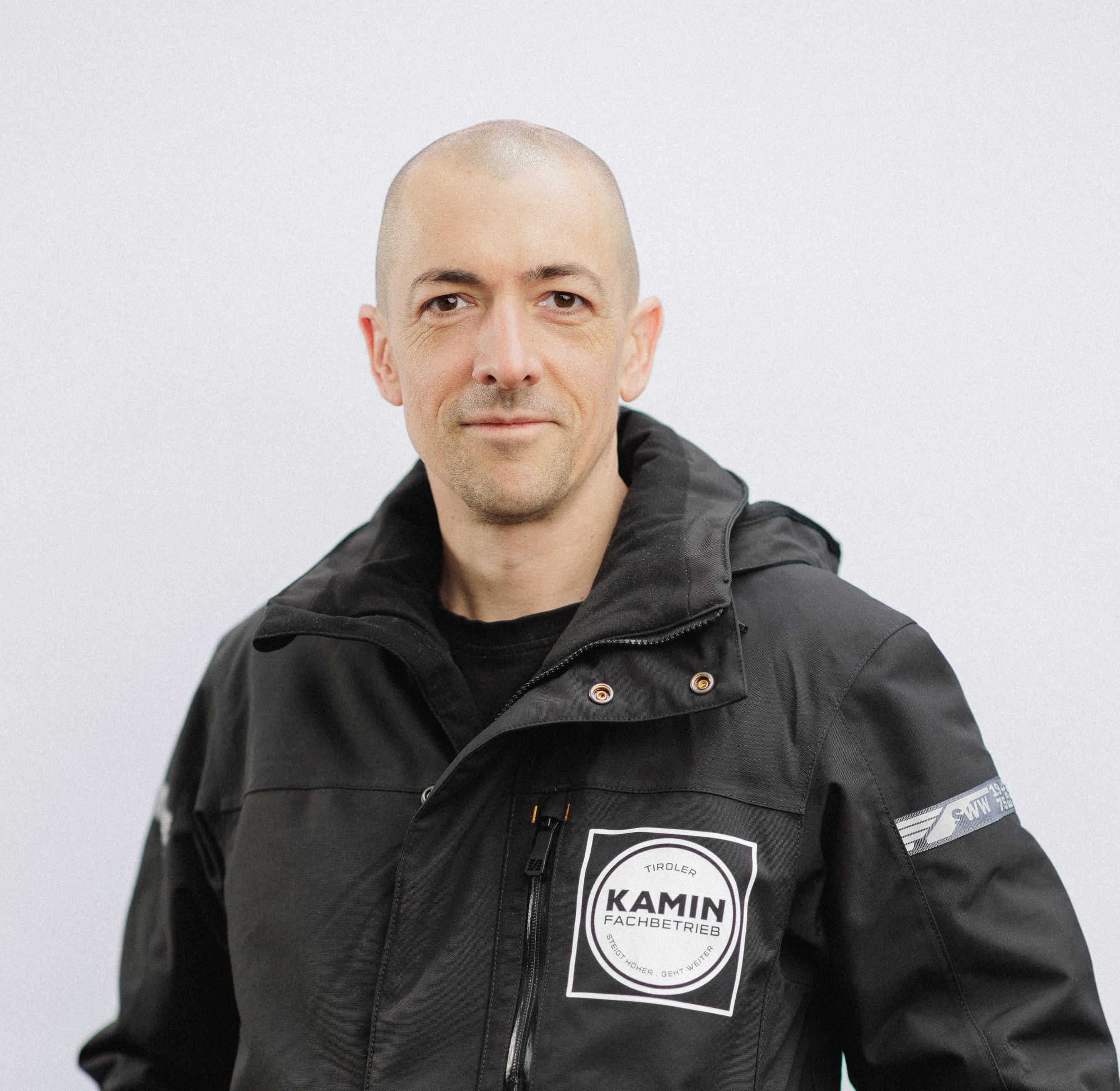 Porträt eines Mannes mit glattem Kopf und schwarzer Jacke mit Kamin-Fachbetrieb-Logo auf weißem Hintergrund.