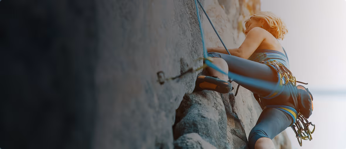 Frau klettert an einer Felswand, gesichert mit Seilen und Kletterausrüstung.