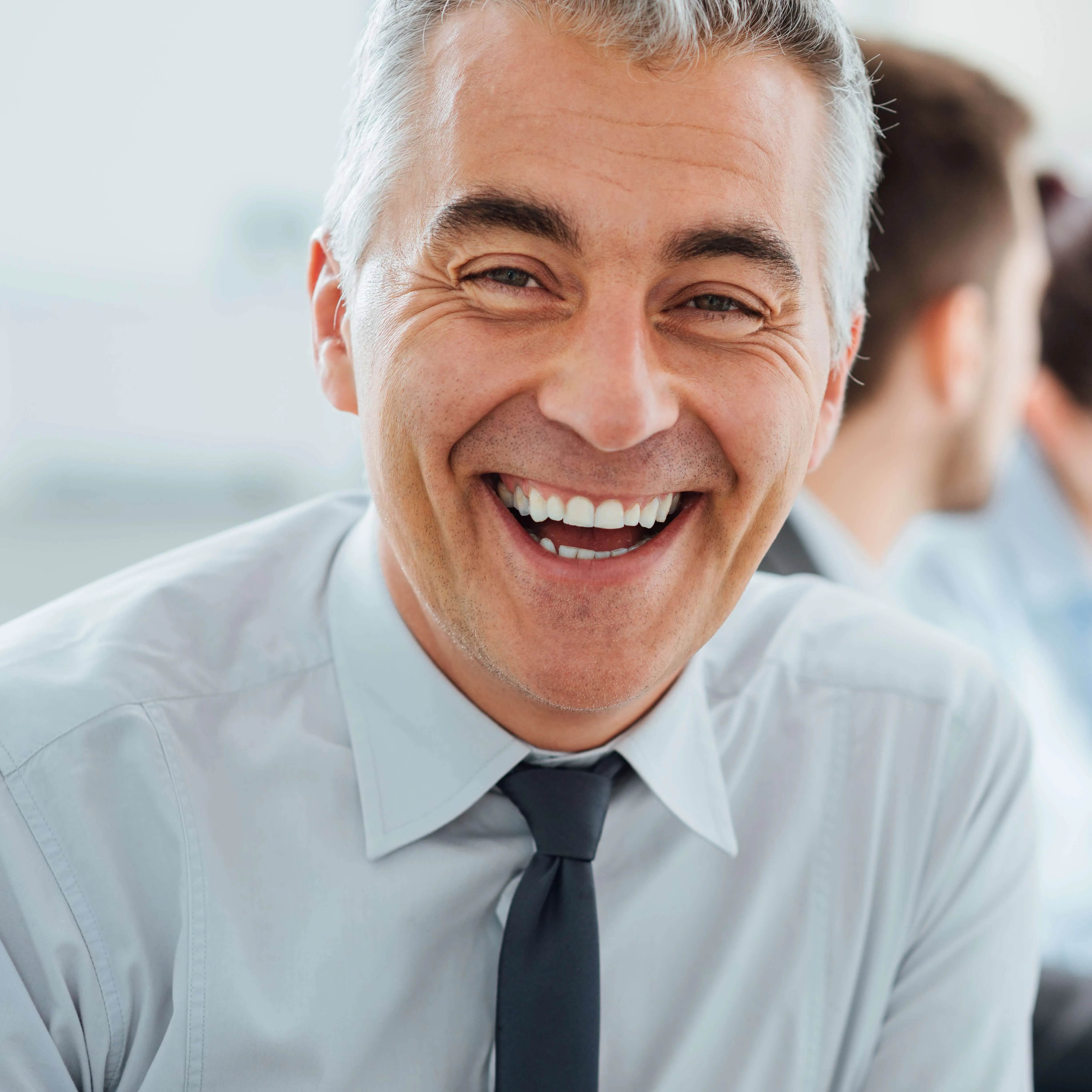 Close-up of a smiling middle-aged man with grey hair, wearing a light shirt and tie, looking directly at the camera in an office setting.