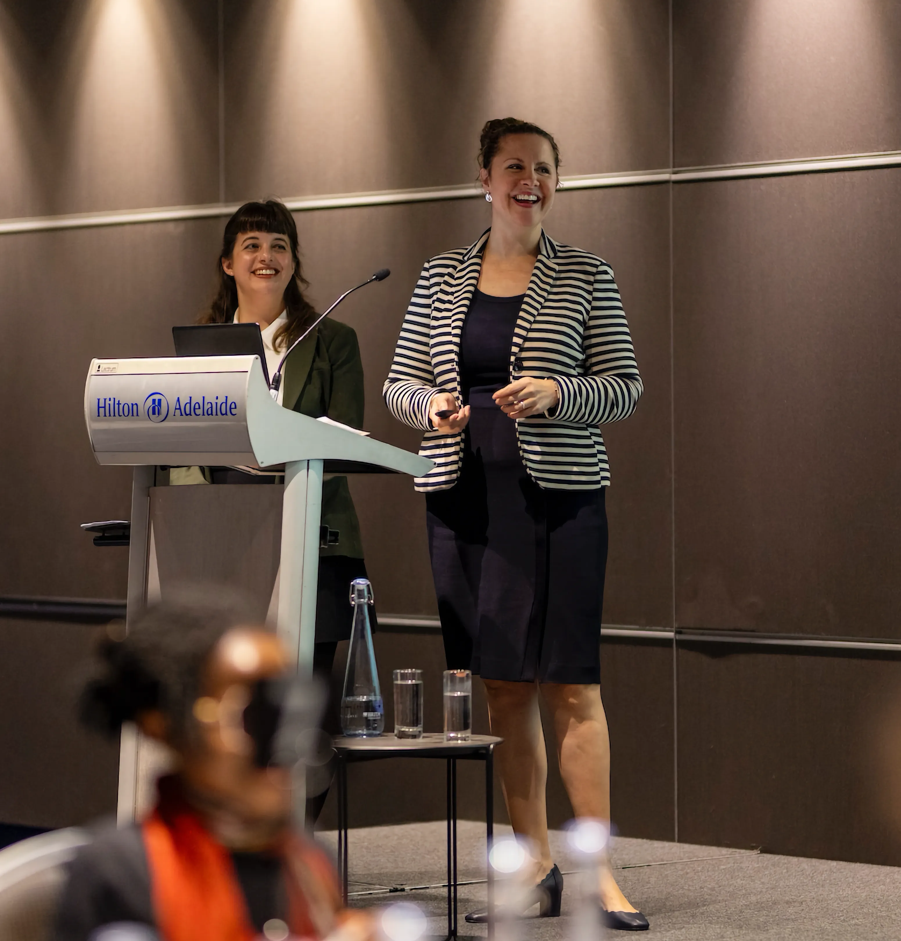 Two women presenting at a podium in a conference room, smiling as they address an audience, with one holding a remote and the other standing behind a laptop.
