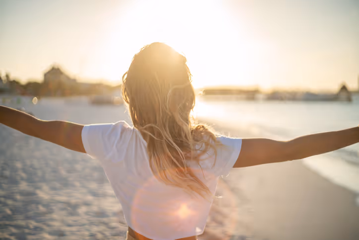 Woman with arms outstretched facing the sun on a sandy beach at sunset.