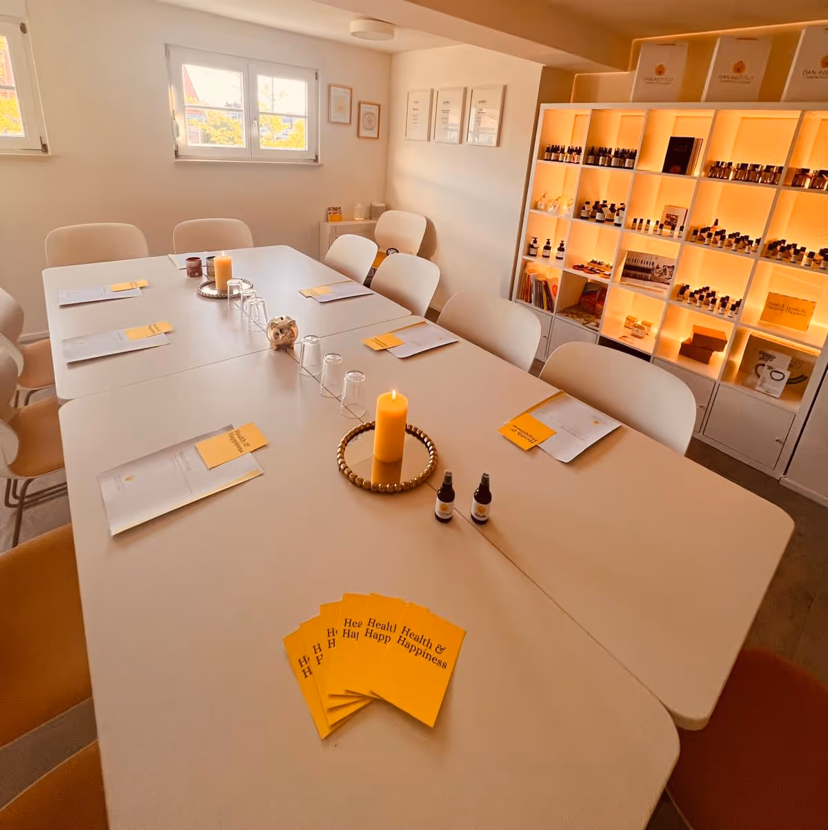 Bright room with long white tables arranged for a meeting, white chairs, lit candle centerpiece, brochures, and a lighted shelf with bottles and products.