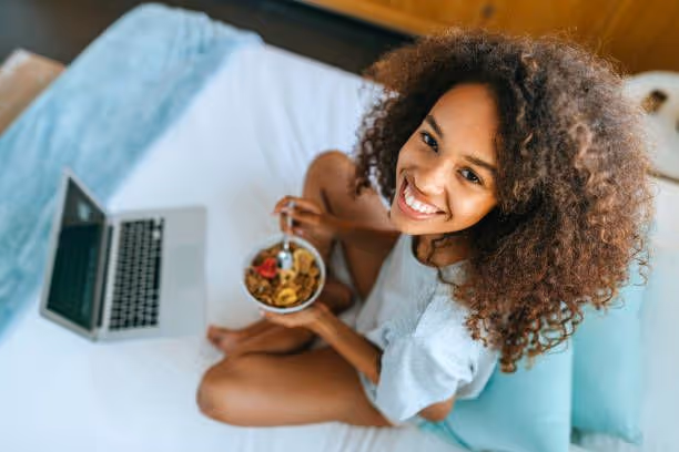 Smiling woman with curly hair sitting on a bed eating cereal from a bowl with a spoon next to an open laptop.