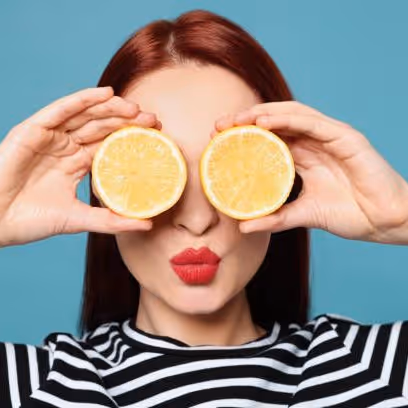 Woman with red hair and red lipstick holding two lemon halves in front of her eyes against a blue background.