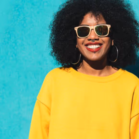 Smiling woman with curly hair, yellow sweater, sunglasses, hoop earrings, and facial piercings against blue background.