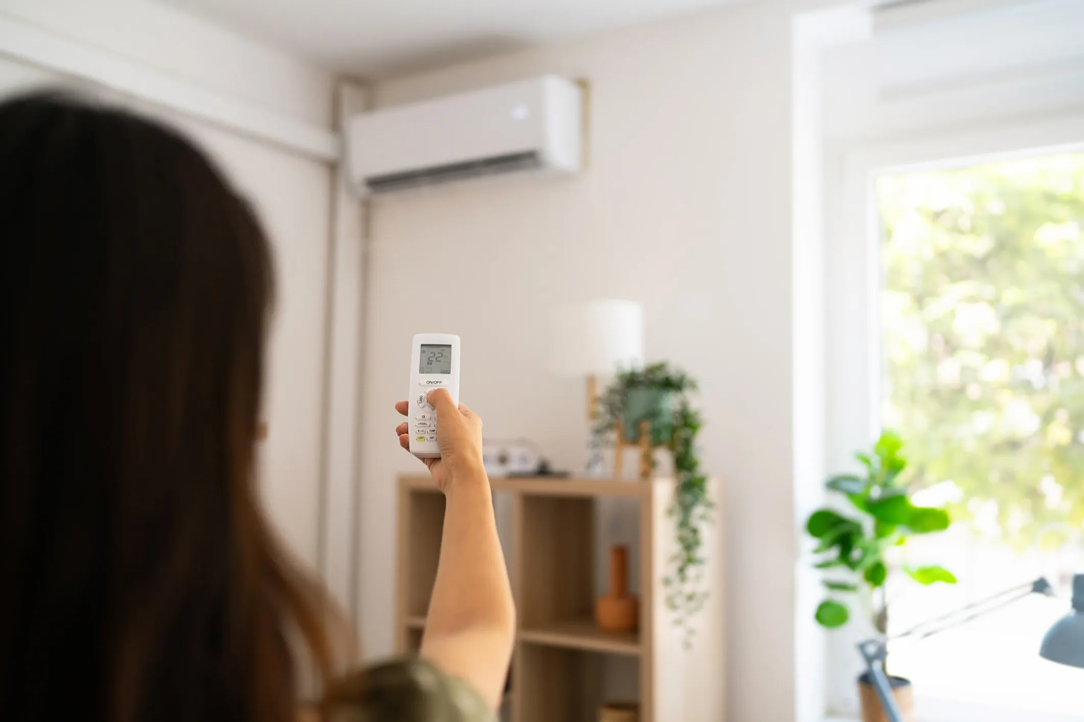  From behind, a person aims a white remote control at a white wall-mounted air conditioning unit in a bright room with a large window and green plants.