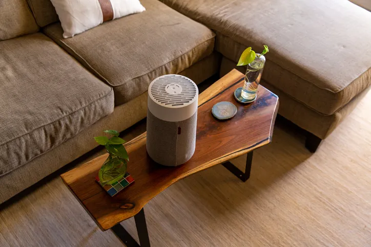 A cylindrical white and grey air purifier with a textured fabric base sits on a live-edge wooden coffee table next to a brown sectional sofa.