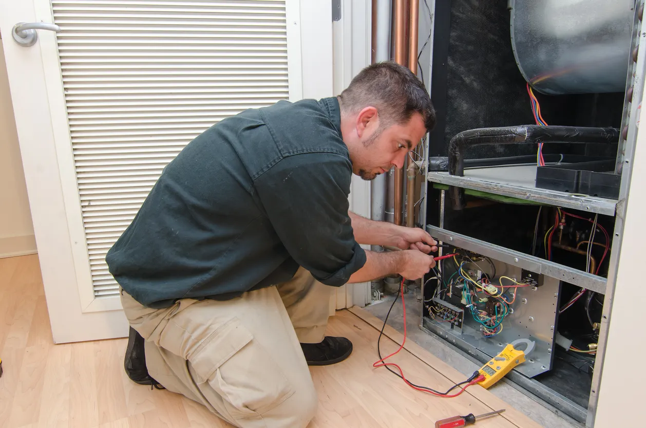 A male technician kneels on a light wooden floor, focused on repairing the internal wiring and components of an open HVAC unit, with a yellow multimeter connected.