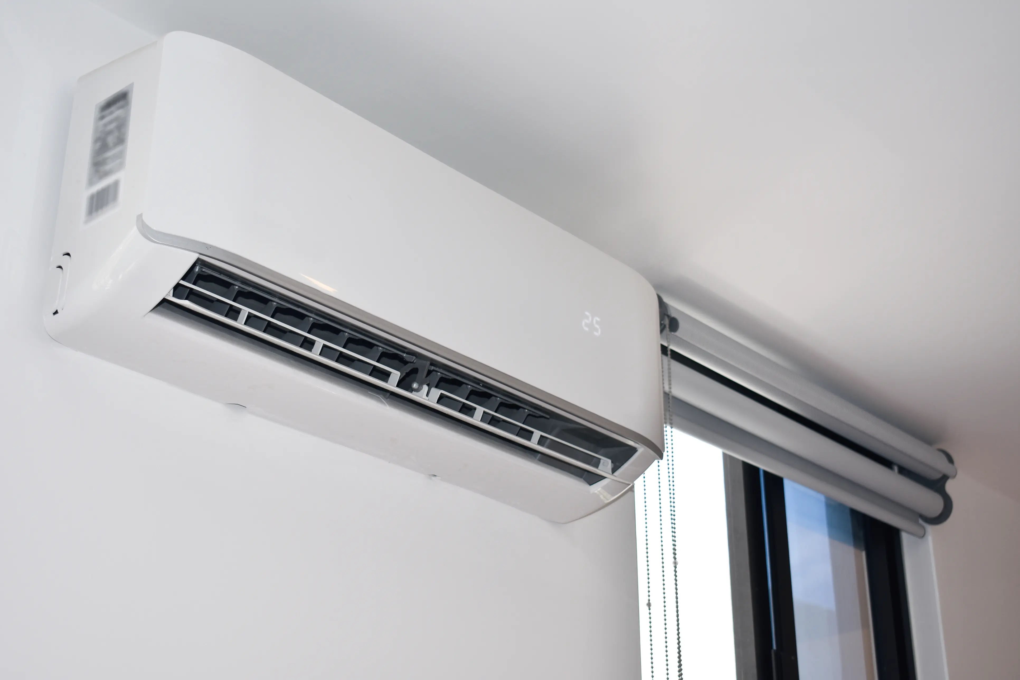 A white ductless mini-split air conditioning unit is mounted high on a white wall, displaying "25" on its digital screen, with a window and roller blind visible on the right.