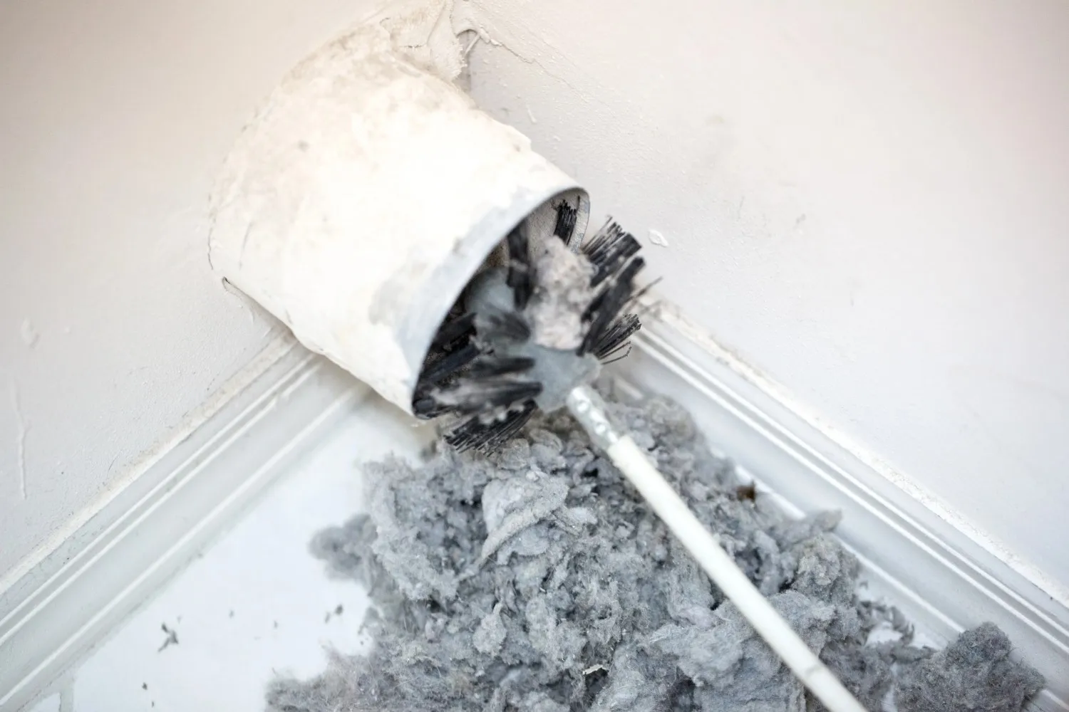  A dryer vent brush is extracting a large pile of gray lint from a white dryer vent pipe opening in a corner.