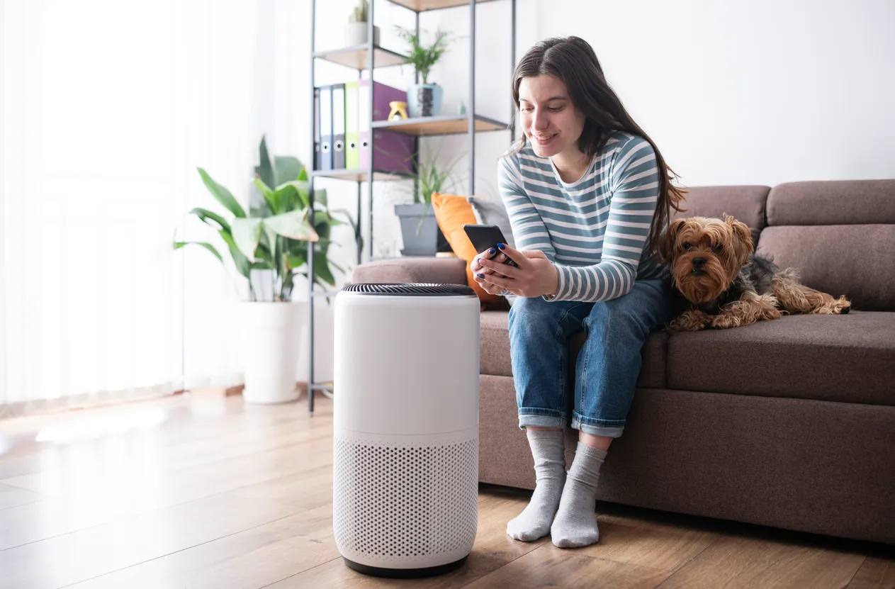 A women sitting next to a air purifier using a phone in a sofa with her dog
