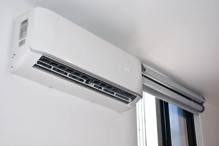 A white mini-split air conditioner unit mounted on a white wall next to a window with blinds.