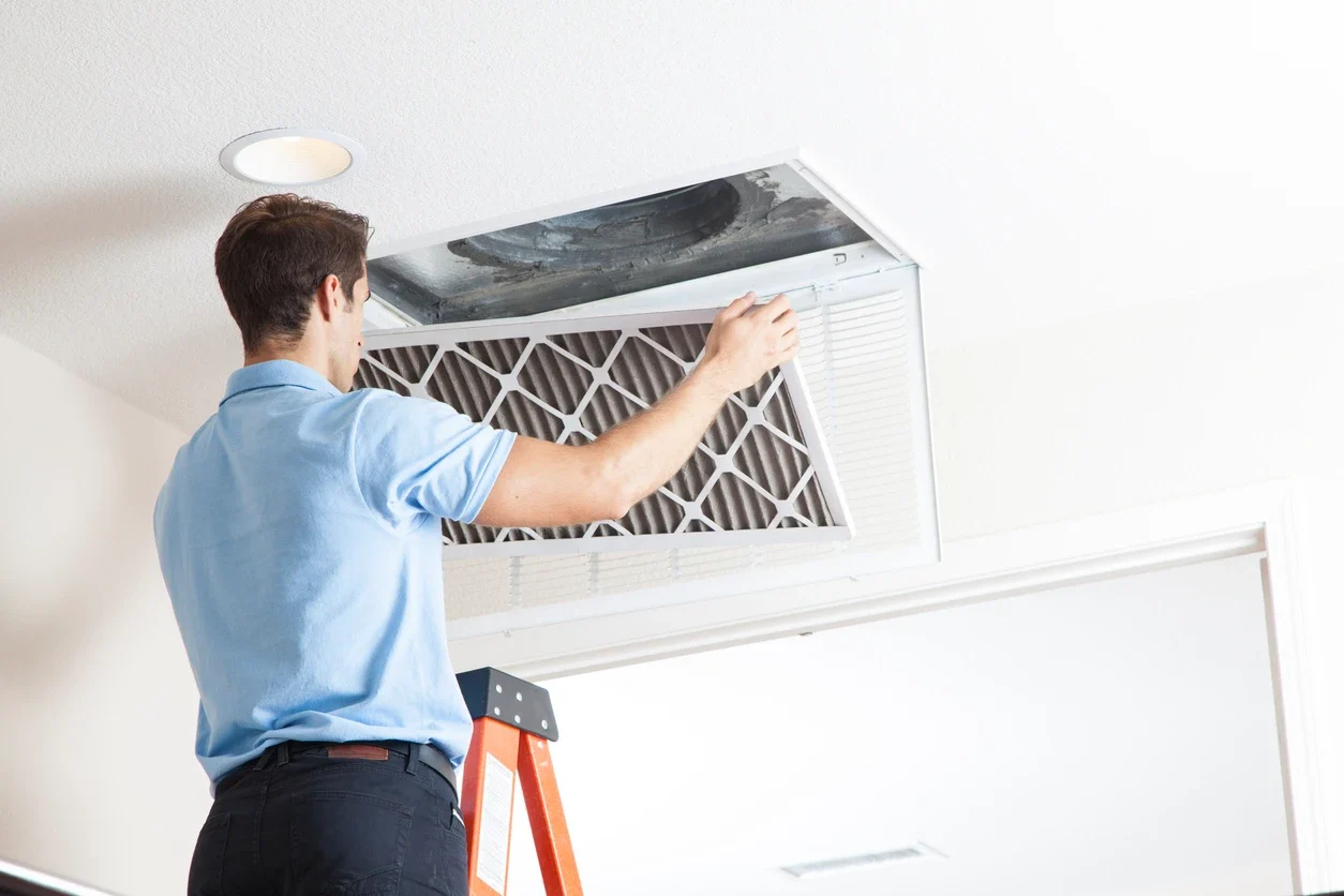 A service technician on a ladder is replacing a dirty air filter in a home's ceiling air vent.