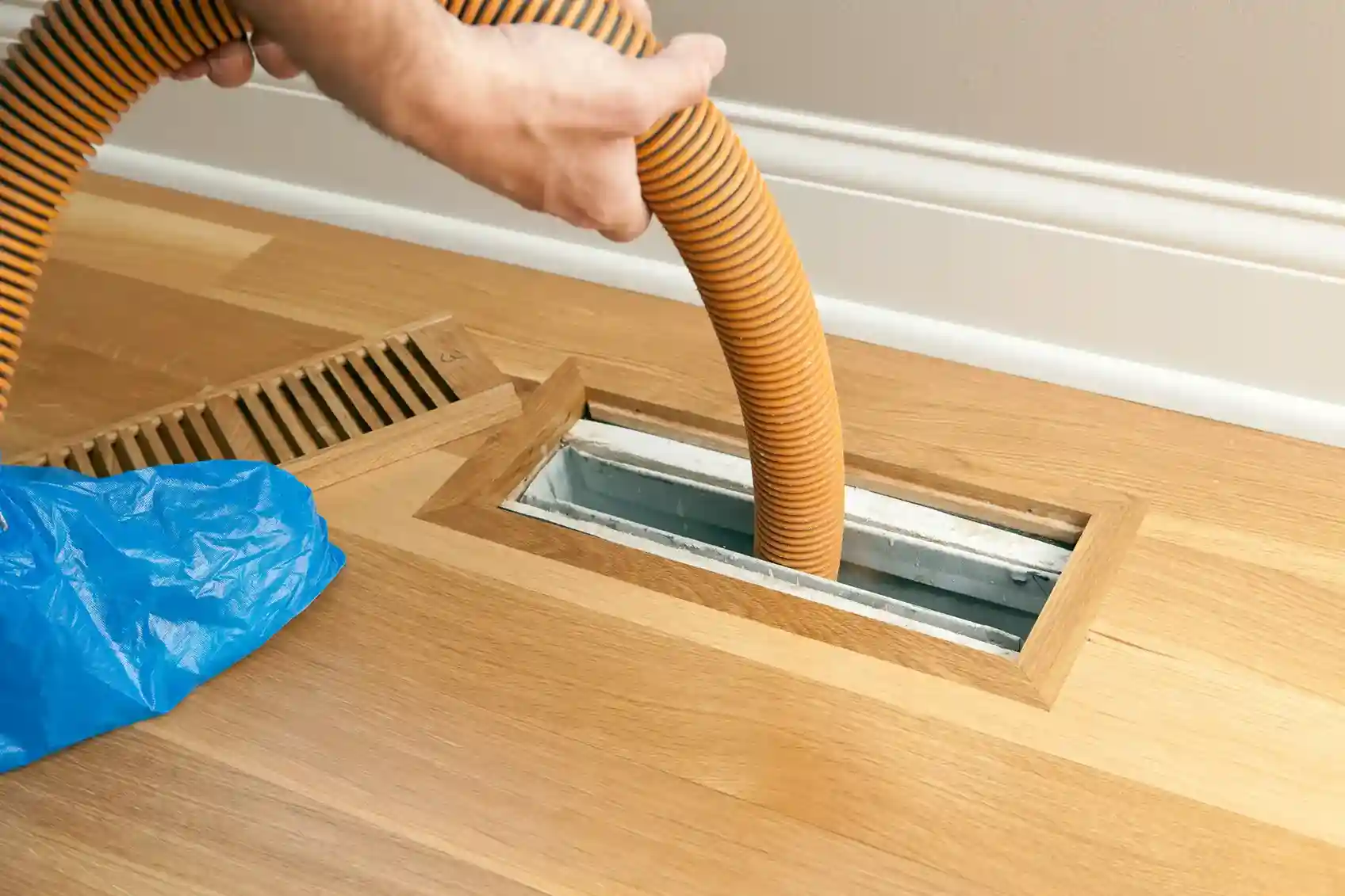 A person holds a striped orange and yellow vacuum hose, inserting it into a floor vent on a light wood floor next to a blue bag of discarded materials.