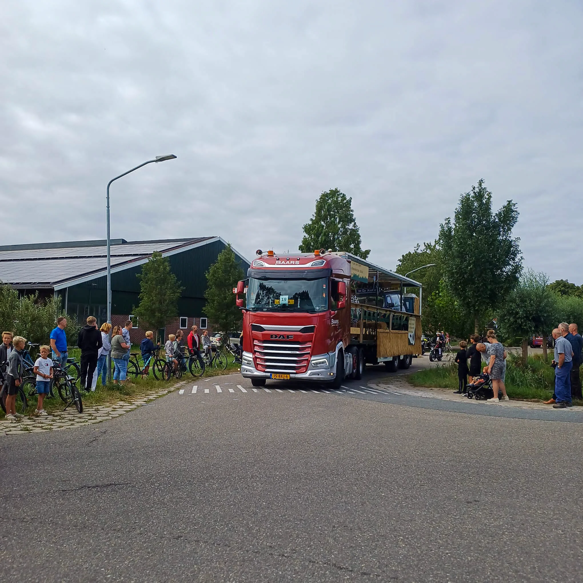 ##   Een rode vrachtwagen van Baars Transport trekt een feestwagen door het dorp tijdens de feestweek in Nieuwland, omringd door toeschouwers.