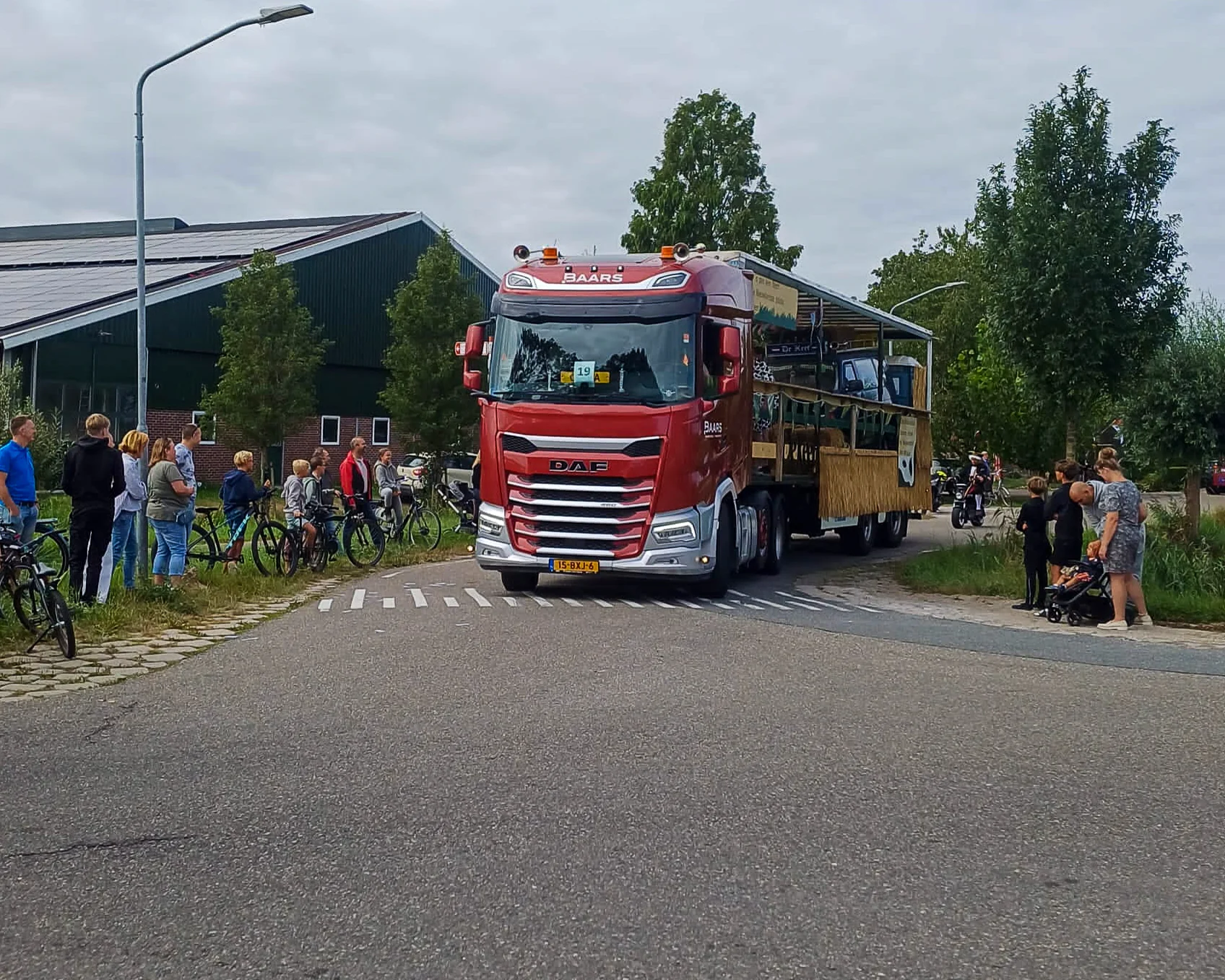 ##   Een rode vrachtwagen van Baars Transport trekt een feestwagen door het dorp tijdens de feestweek in Nieuwland, omringd door toeschouwers.