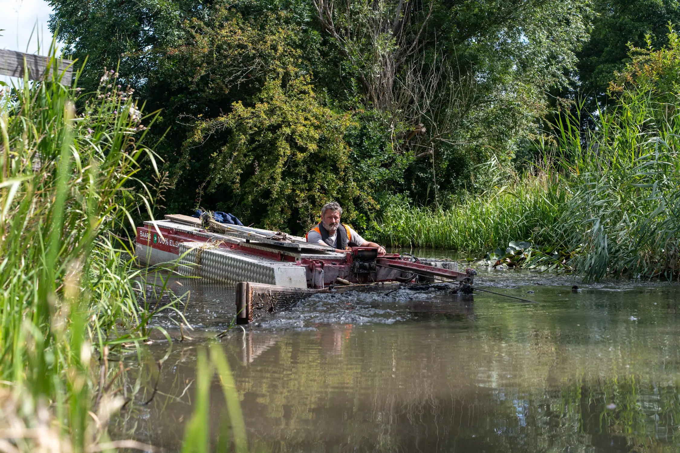 Baars medewerker stuurt elektrische baggerboot door smalle watergang om waterplanten te verwijderen