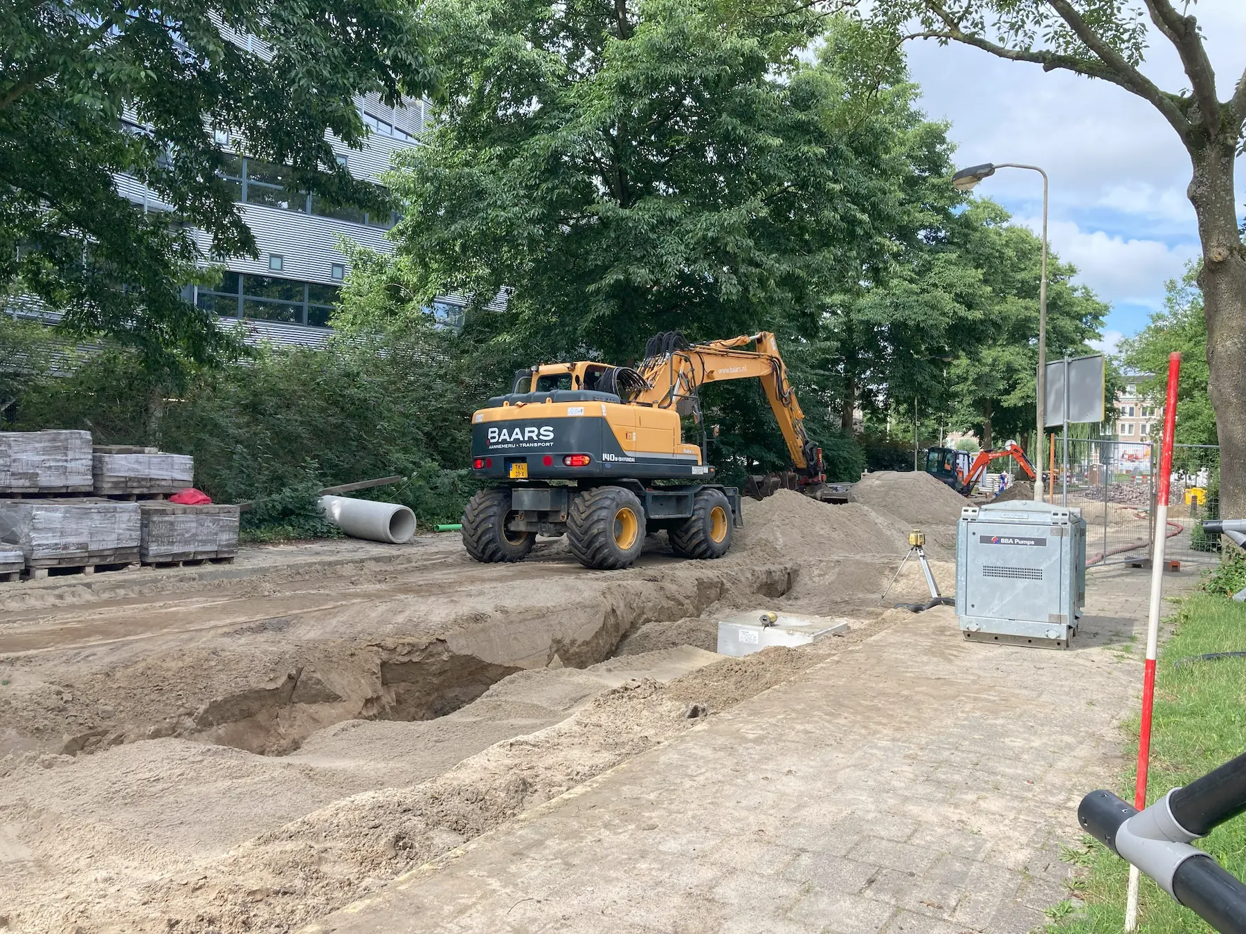 Straatbeeld met oranje minigraver, werklieden en open sleuven tijdens het rioolproject van Baars in de Gildenwijk.