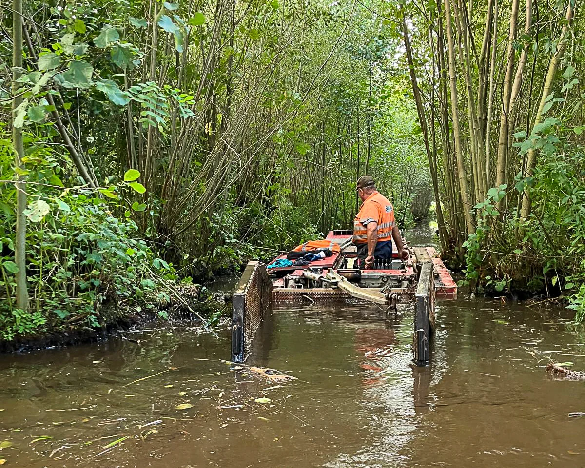 werknemer van Baars bedient een kleine baggerboot in een smalle, dichtbegroeide watergang, omringd door struiken en bomen.