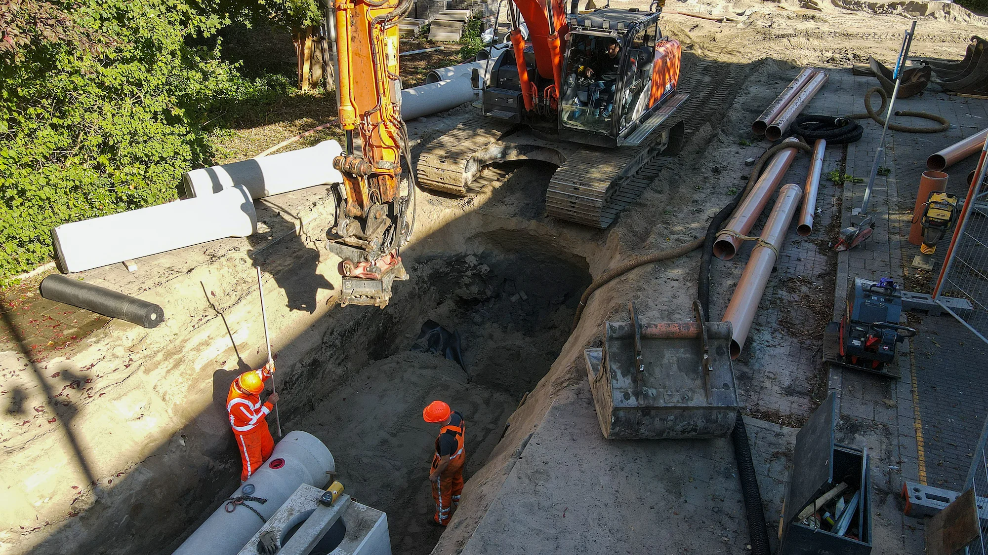 Twee grondwerkers begeleiden het plaatsen van een betonnen rioolbuis terwijl een graafmachine de buis laat zakken tijdens werkzaamheden in de Gildenwijk