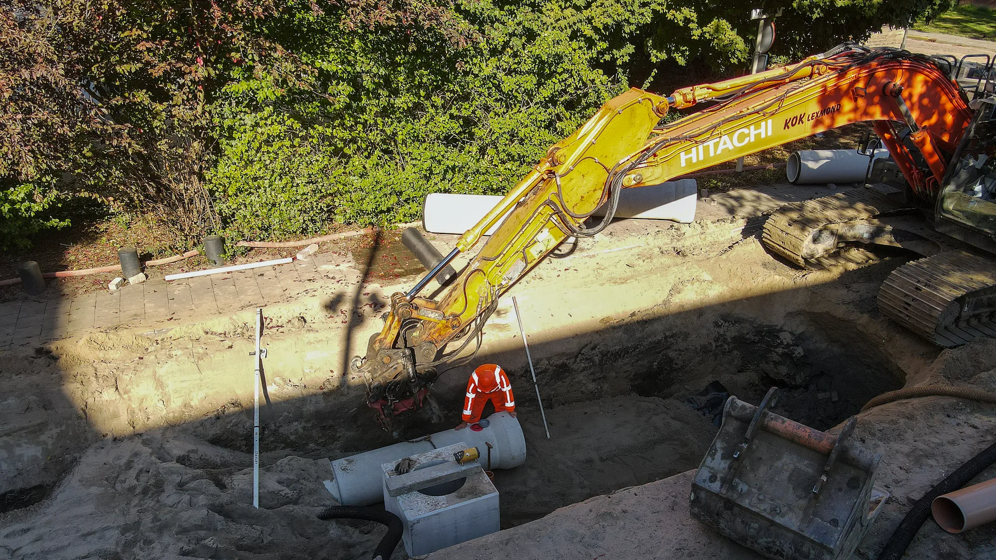 Grondwerker in een diepe sleuf plaatst betonnen rioolbuis met ondersteuning van een graafmachine tijdens rioolreconstructie in een woonwijk