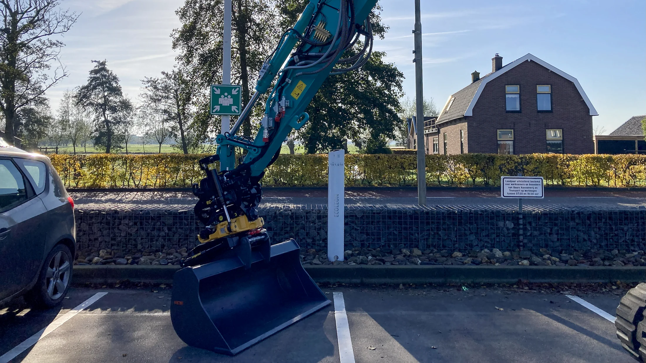 Detail van de voorzijde van de midigraver met maaikorf en gripper, met op de achtergrond het landelijke uitzicht bij Baars.