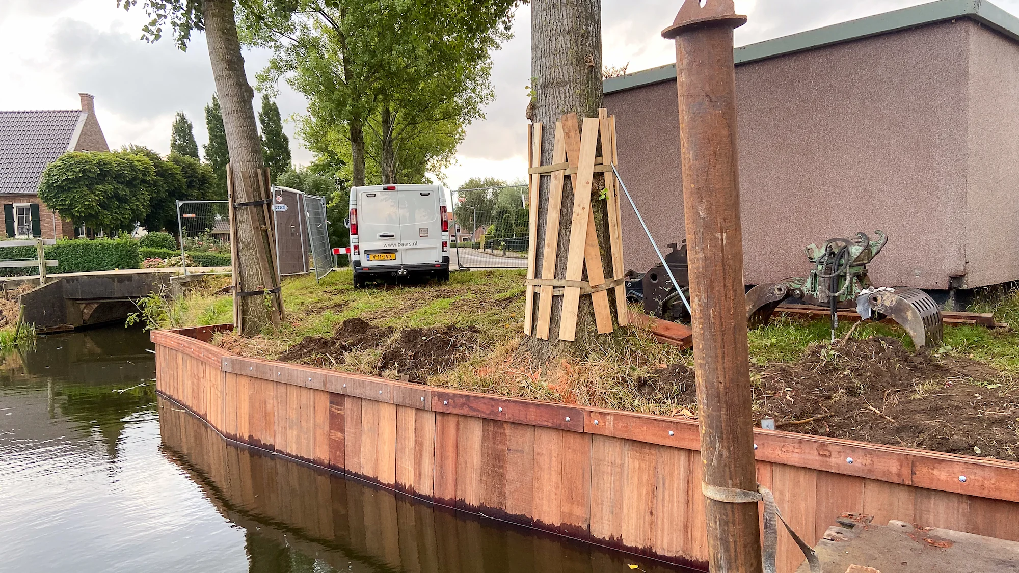 Nieuw geplaatste hardhouten damwand langs de waterkant, met boomverankering en bouwmaterialen op de oever.