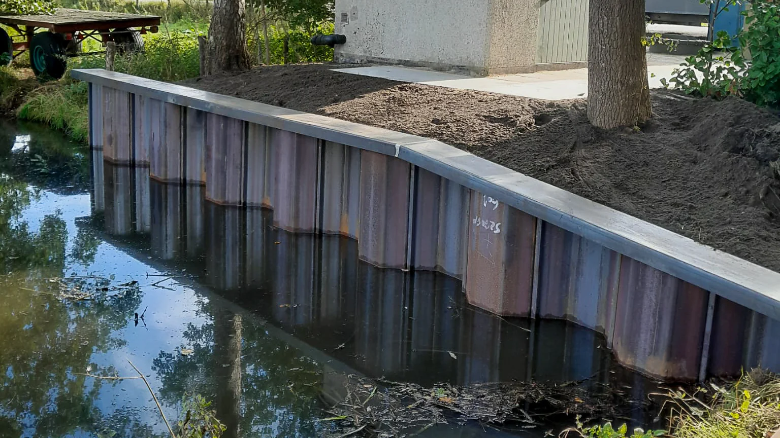 Stalen damwandconstructie met nette afwerking, geplaatst langs een watergang bij een betonnen gebouw in een groene omgeving.