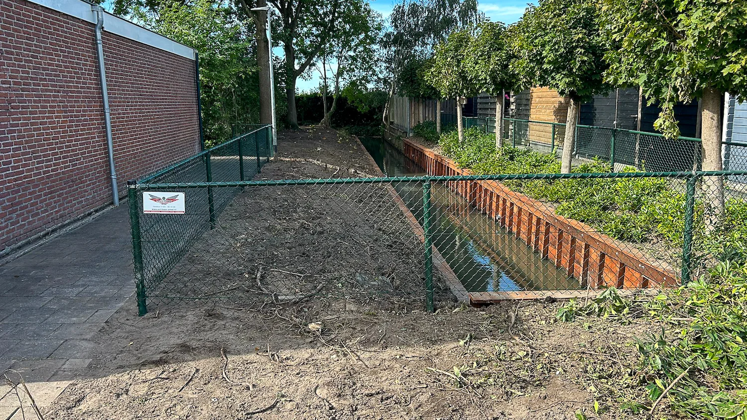 Net afgewerkte hardhouten beschoeiing langs een sloot tussen achtertuinen, met nieuwe afrastering en opgeschoonde berm.