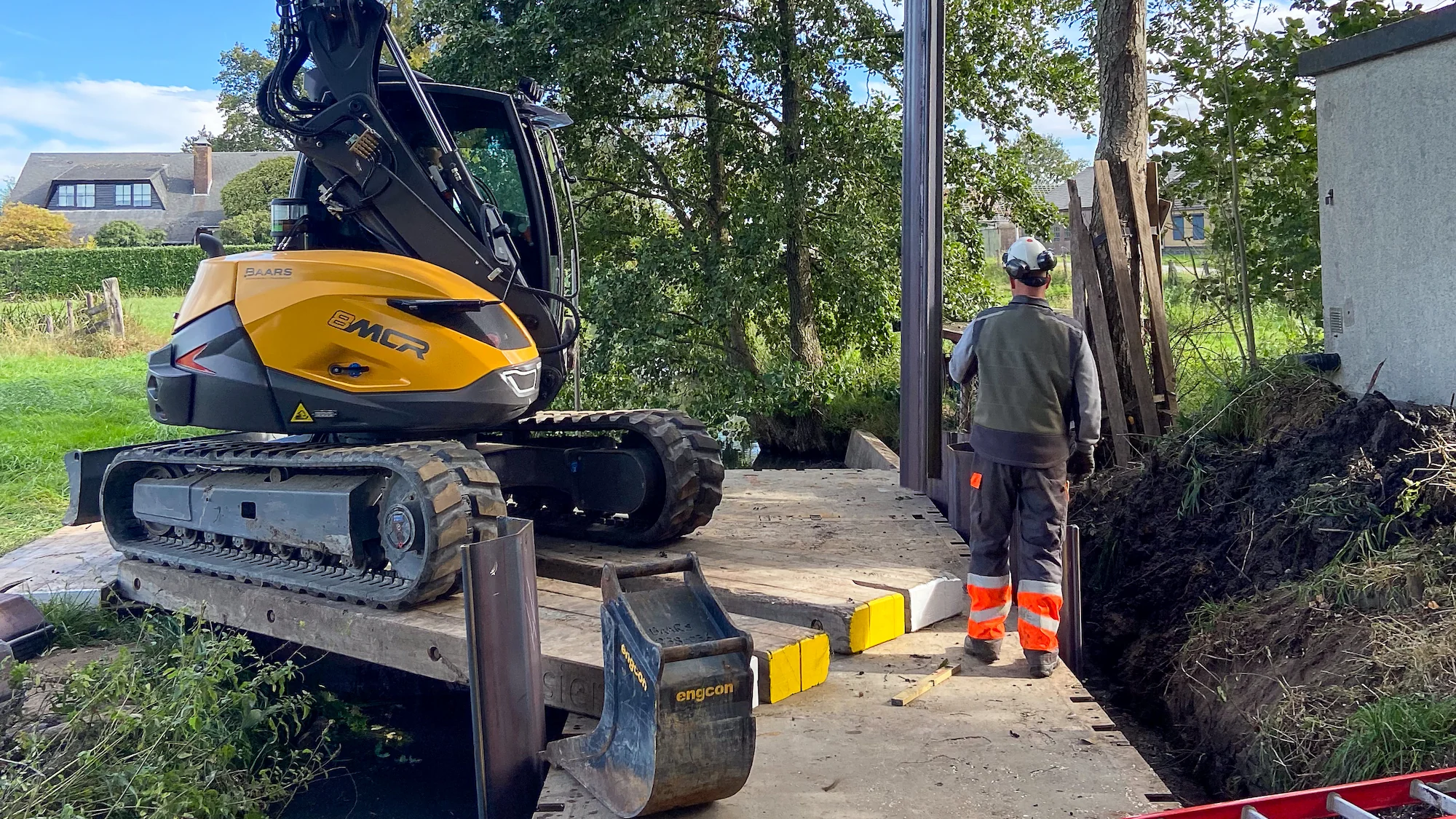 Grondwerker aan het werk met een Mecalac op ponton, bij het plaatsen van een damwand op moeilijk bereikbare locatie aan het water.