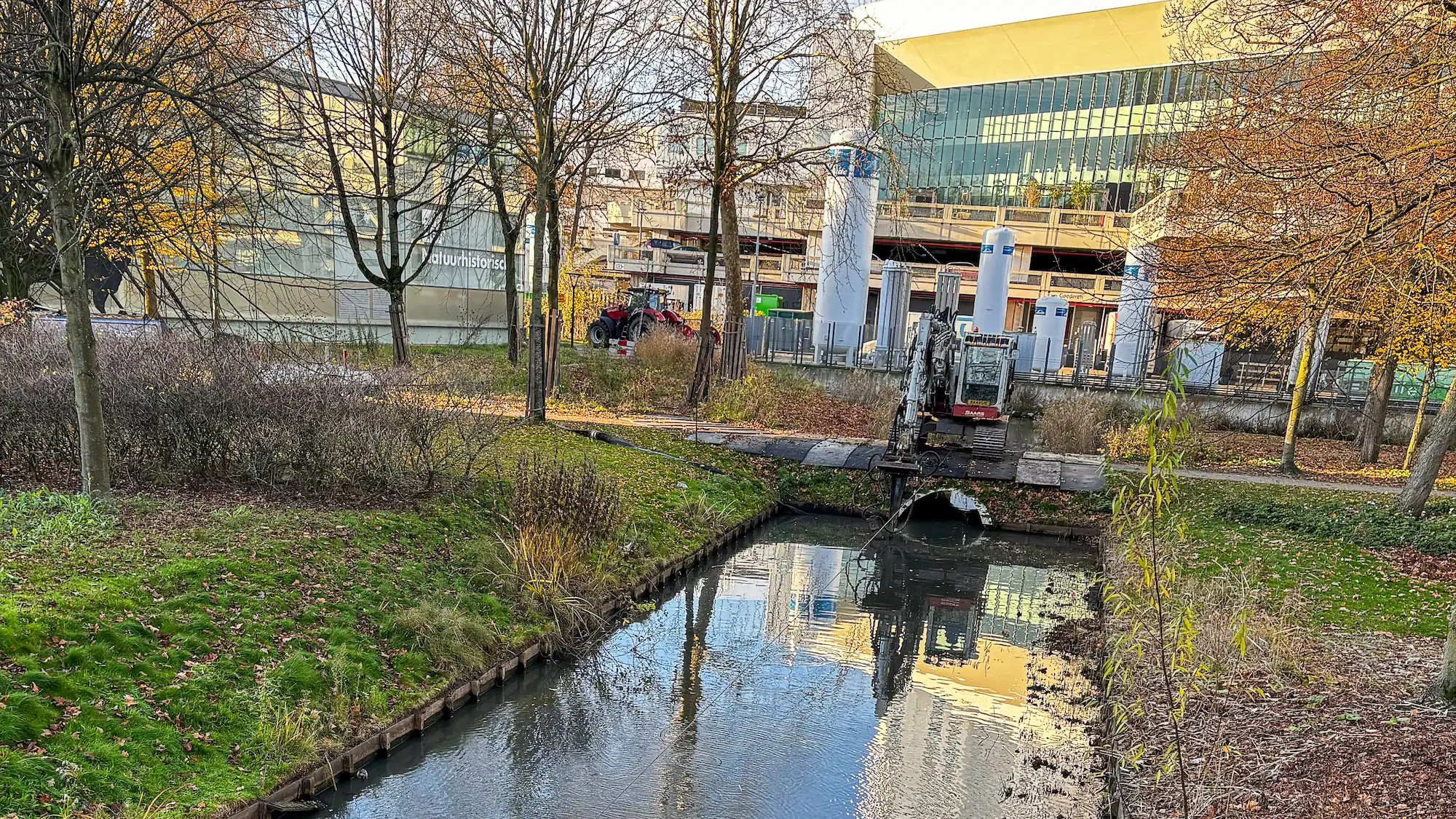 Een minigraver op ponton voert baggerwerkzaamheden uit in het Museumpark in Rotterdam, met het Natuurhistorisch Museum op de achtergrond.