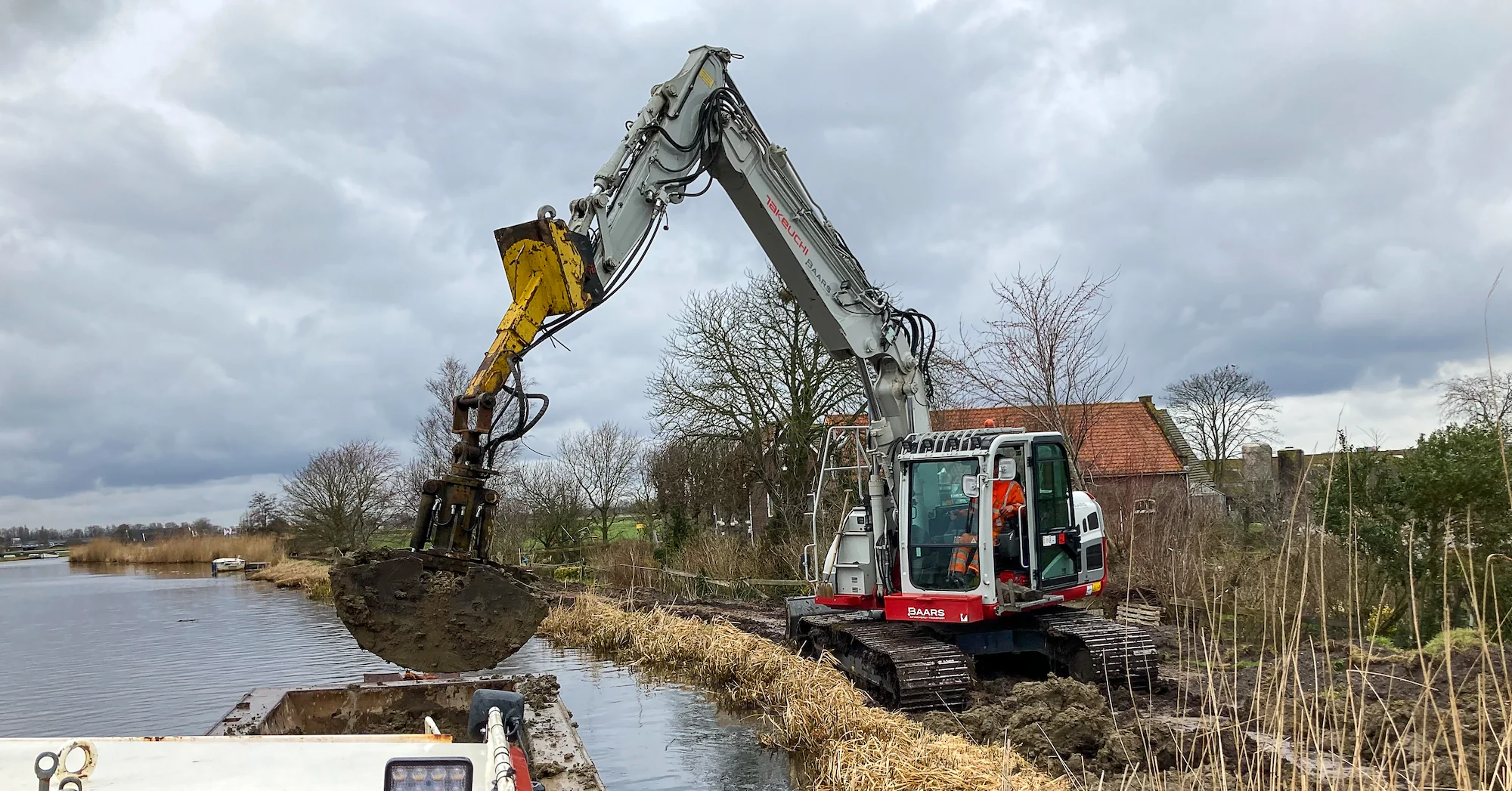 Takeuchi graafmachine plaatst grond in beunbak vanaf de oever