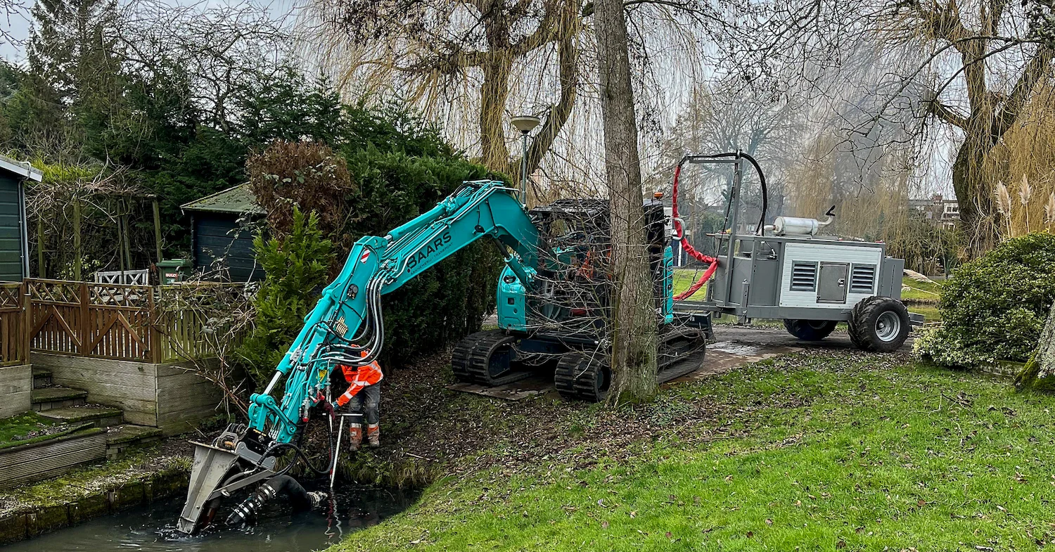 Baars graafmachine met nieuwe powerunit aan het werk bij baggeren van smalle watergang in woonwijk