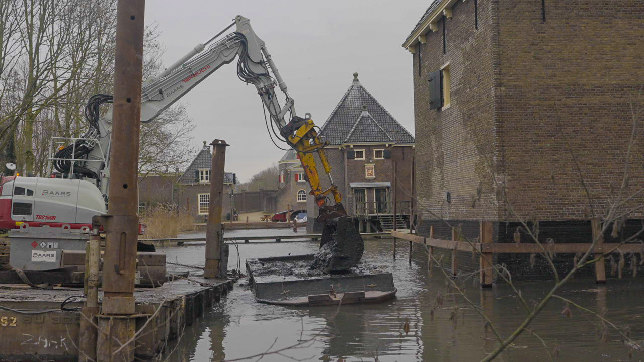 Grote Baars graafmachine werkt vanaf ponton bij baggerwerkzaamheden in watergang nabij historisch Kruithuis