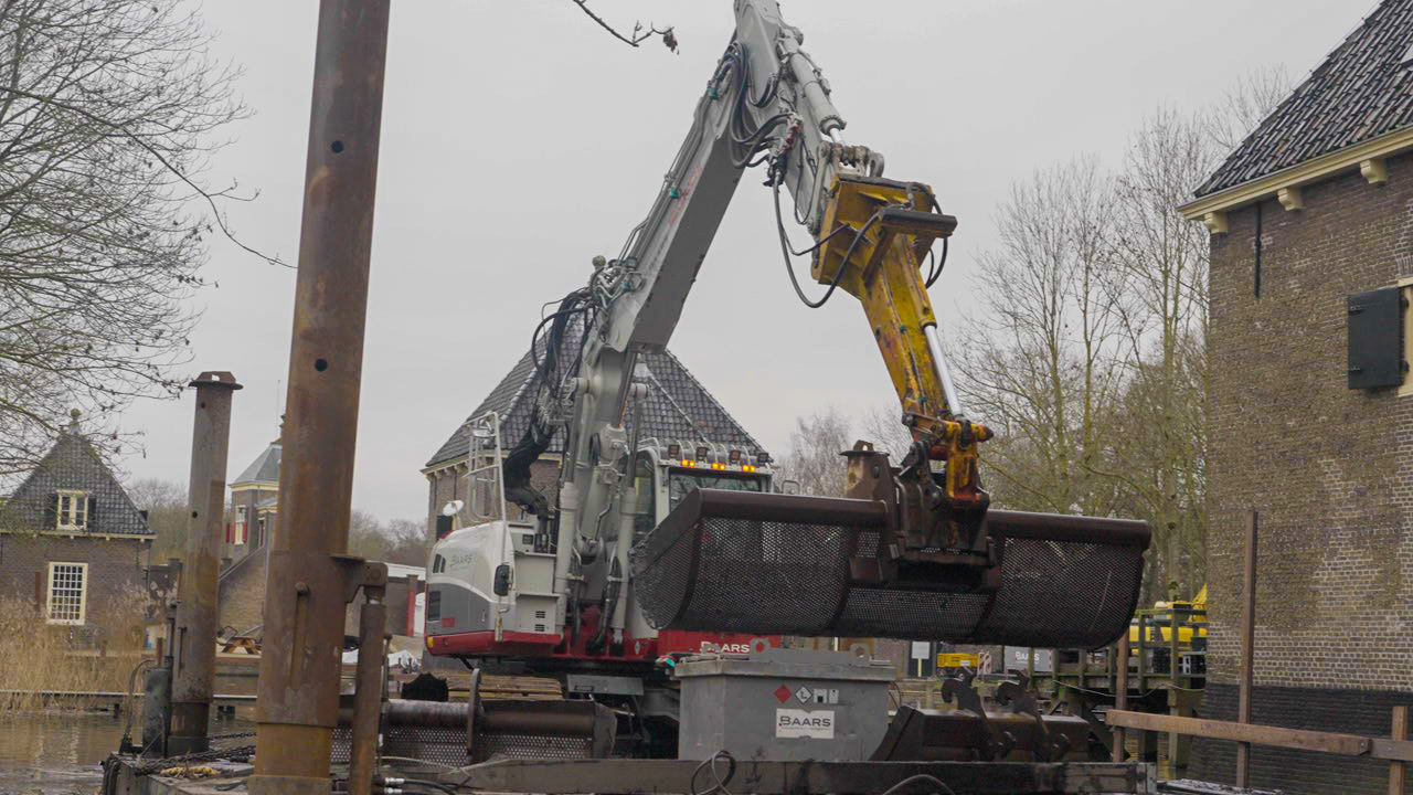 Grote Baars graafmachine werkt vanaf ponton bij baggerwerkzaamheden in watergang nabij historisch Kruithuis