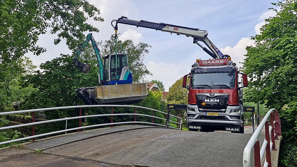 Een vrachtwagen met autolaadkraan hijst een compacte minifloat over een brug naar een smalle watergang voor werkzaamheden aan beschoeiing en baggerwerk.