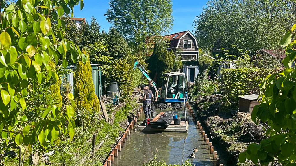 Medewerkers van Baars vervangen beschoeiing langs een smalle watergang in een volkstuincomplex. Op de achtergrond staat een compacte minifloatkraan tussen het groen en de tuinen.