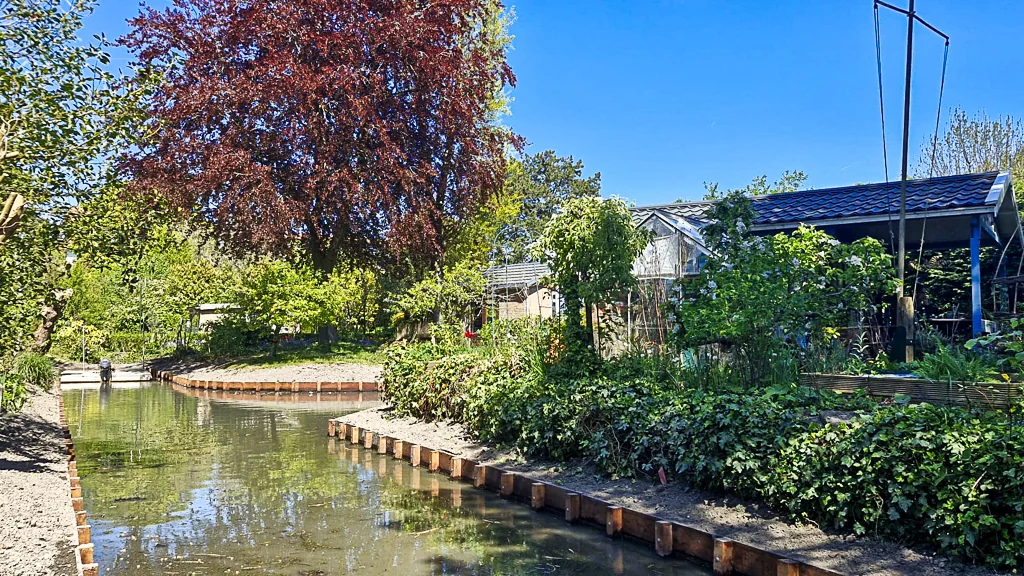 Overzicht van een vernieuwde watergang in een volkstuincomplex met nieuwe beschoeiing, groene oevers en een nette afwerking van het project.