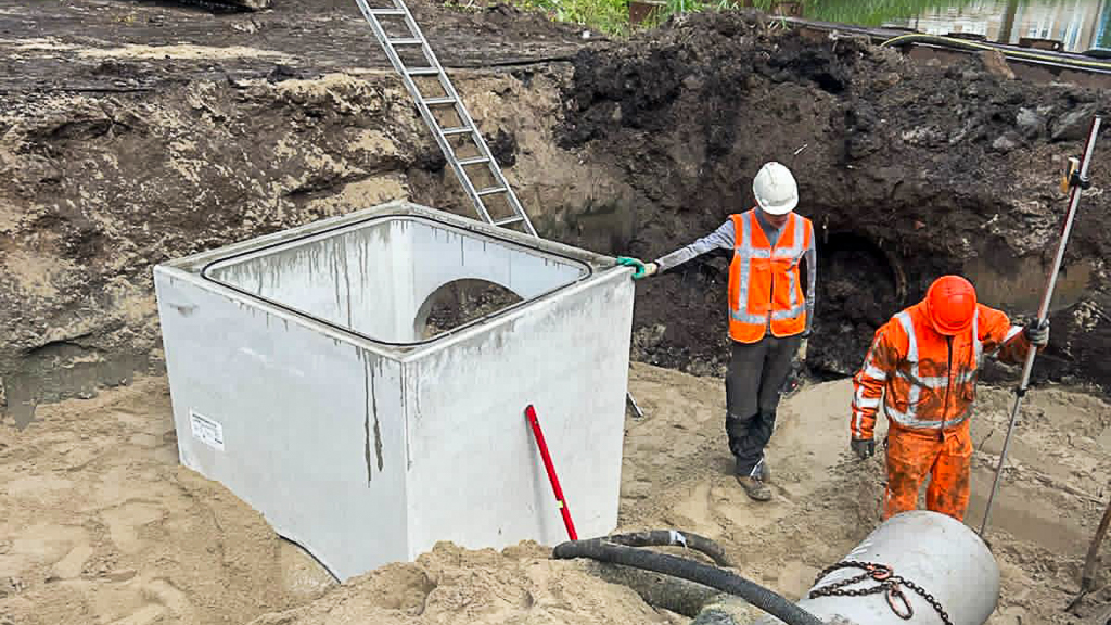 Werkzaamheden aan een nieuwe rioolput nabij een watergang. Medewerkers bereiden de aansluiting van de betonnen elementen voor in een zorgvuldig uitgegraven bouwput.