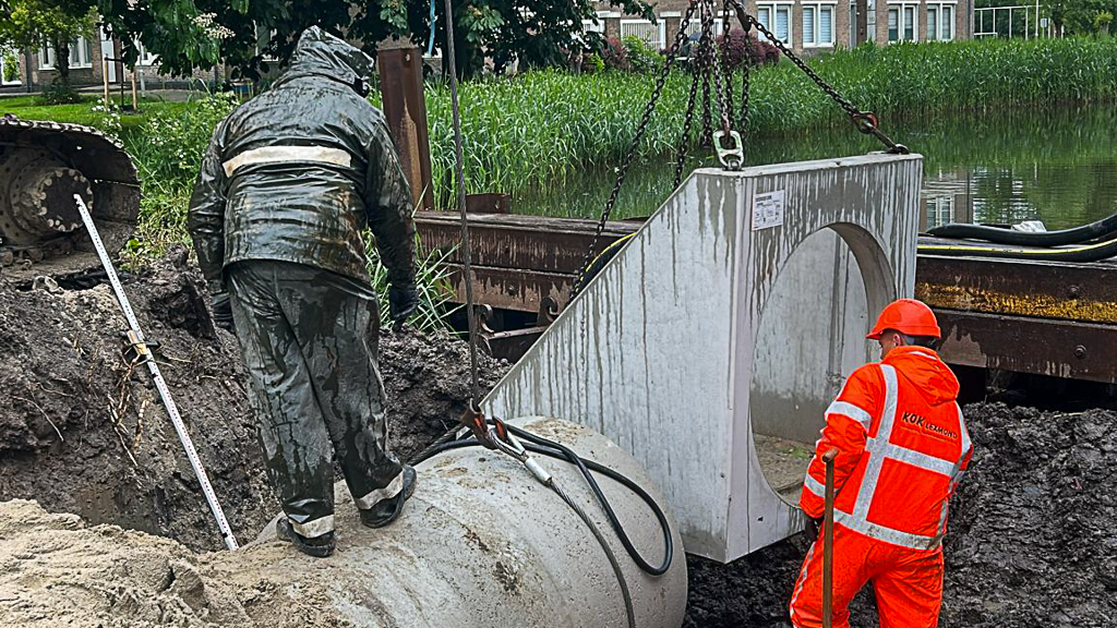 Plaatsen van een betonnen duikerelement tijdens rioolwerkzaamheden. Een medewerker controleert de hoogte en positie terwijl de kraan het element nauwkeurig in de sleuf laat zakken.