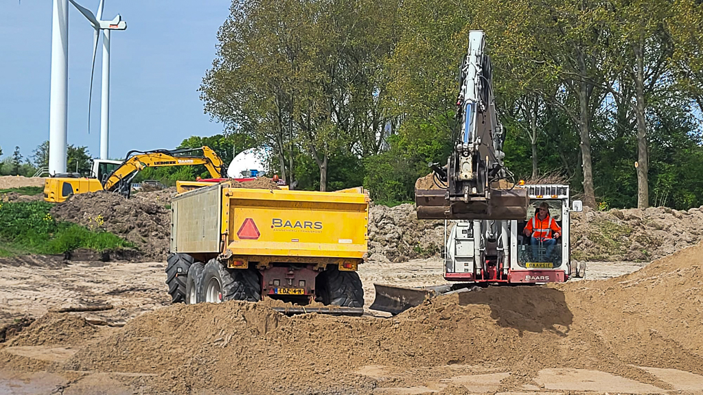 Een machinist laadt grond in een dumper tijdens de aanleg van de nieuwe depotkade. Op de achtergrond zijn aanvullende grondwerkzaamheden zichtbaar.