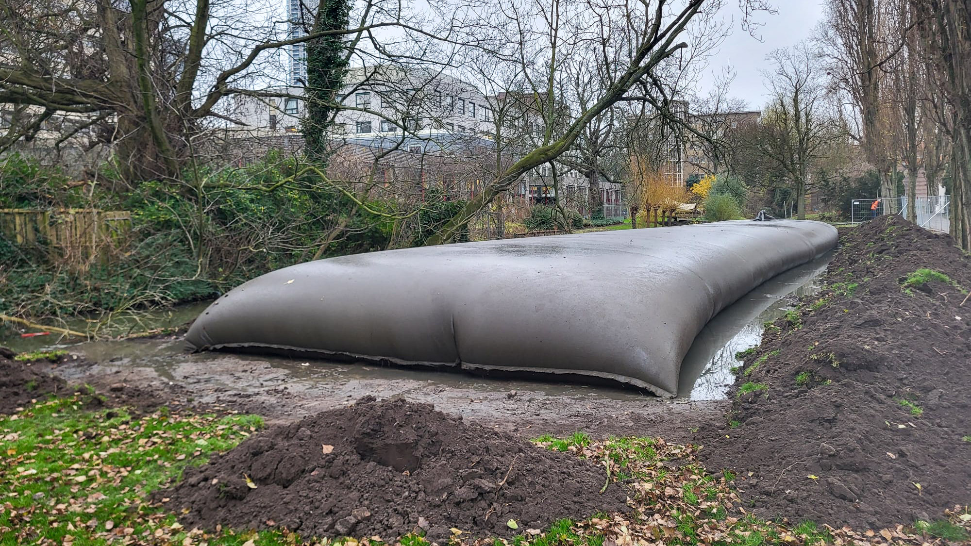 Grote geotextiele slibzak (geotube) gevuld en gelegd in een watergang tussen kale bomen, met grond en bouwhekken op de oever.