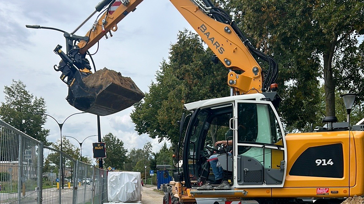 Close-up achteraanzicht van de Liebherr 914 met het Baars-logo, kenteken T-59-HRK en werklampen op de cabine, op een bouwlocatie langs een fietspad.