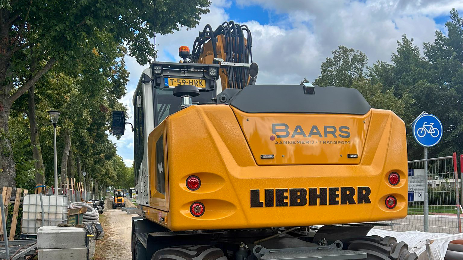 ##   Mobiele graafmachine Liebherr 914 met Baars-logo op de rupsarm, tijdens grondwerkzaamheden aan een straat. De kraan tilt een bak met grond, op de achtergrond bomen en een weg.