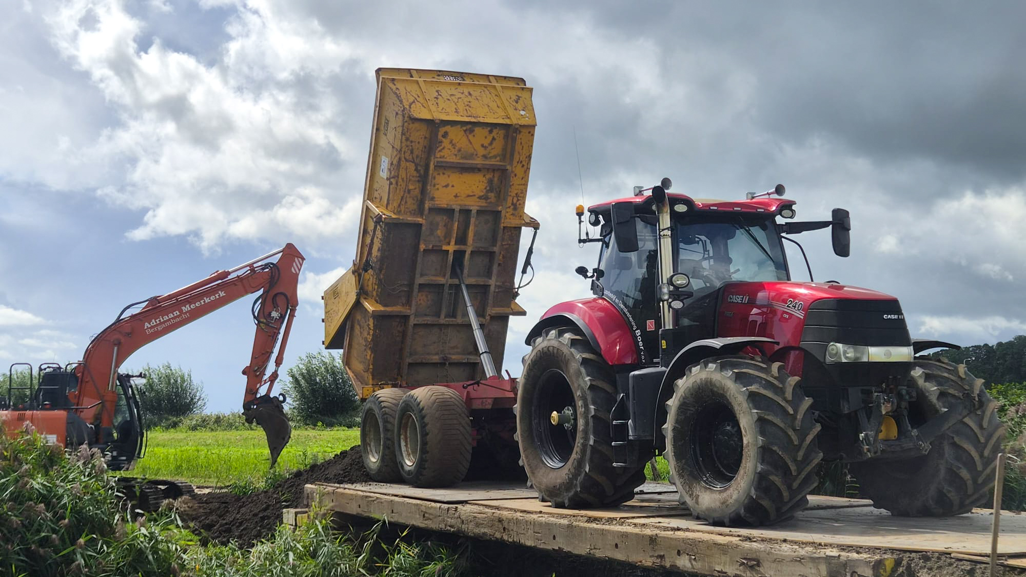 Graafmachine van Adriaan Meerkerk laadt grond in een kipwagen achter een tractor, op rijplaten bij de kadeversterking in Goudriaan.